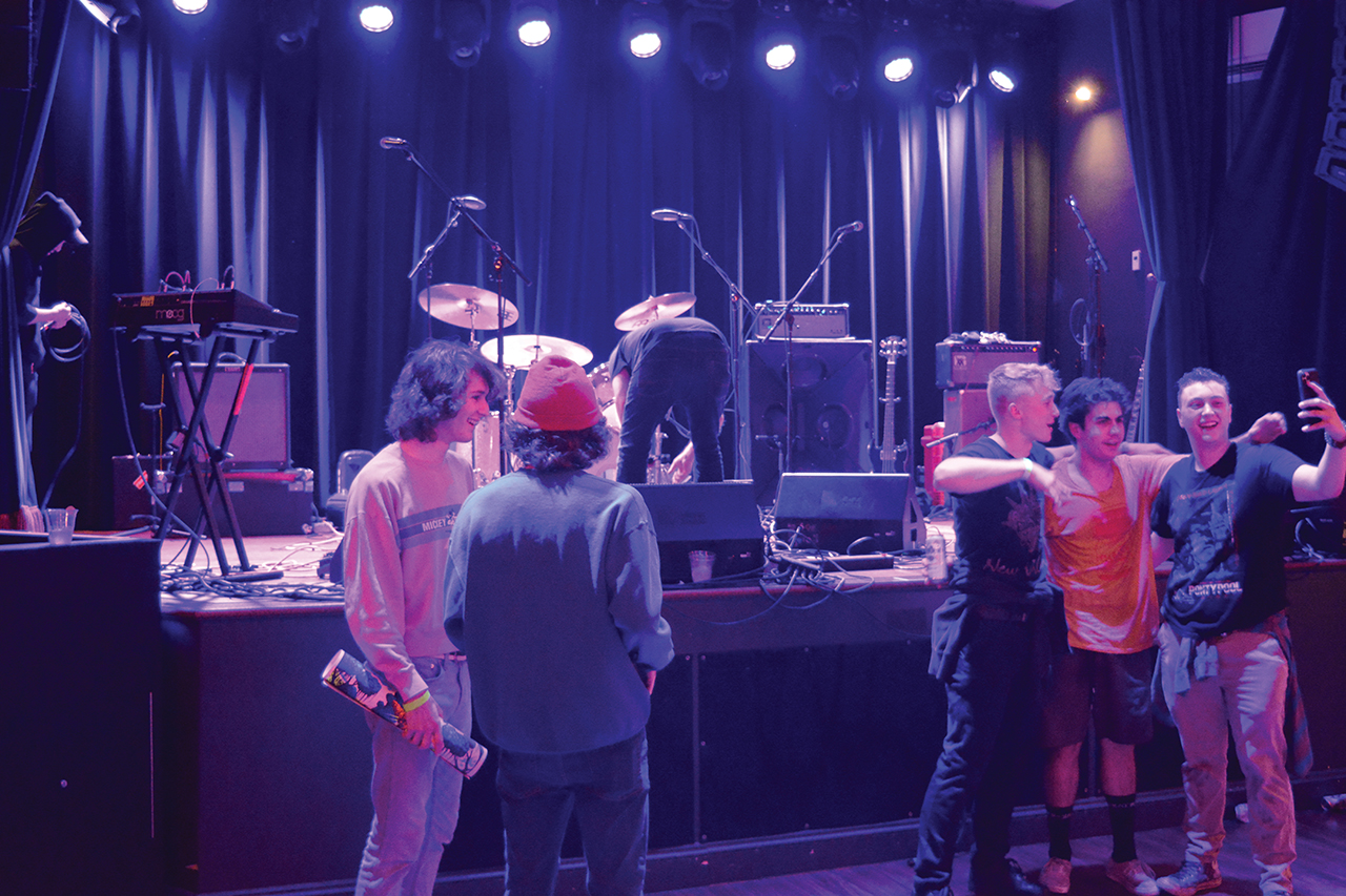 Photo of three people standing in front of the right side of the stage. One raises his arm to take a photo of them with his phone.